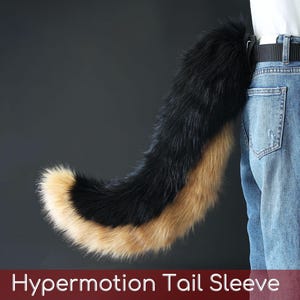 Puede incluir: Una funda de cola de piel sintética negra, marrón y beige sujeta al cinturón de una persona. La cola es curvada y esponjosa, con una punta negra y una parte inferior beige. El texto "Hypermotion Tail Sleeve" está en la parte inferior de la imagen.