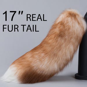 Può includere: Una coda di pelliccia vera lunga 43 cm, con un motivo marrone e bianco. La coda è fissata a un supporto nero.