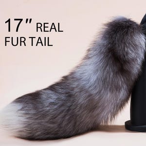 Può includere: Una coda di pelliccia vera lunga 43 cm, di colore grigio chiaro. La coda è fissata a un supporto nero.