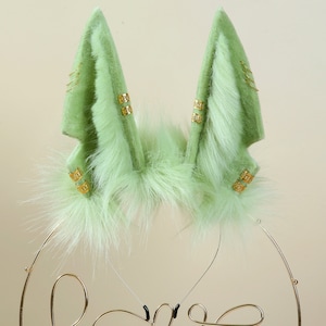 May include: A pair of green faux fur bunny ears with gold accents. The ears are attached to a headband.