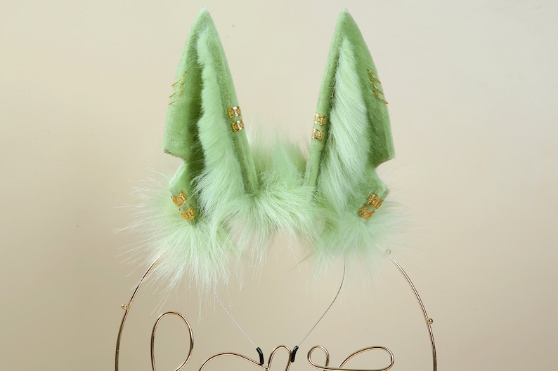 May include: A pair of green faux fur bunny ears with gold accents. The ears are attached to a headband.
