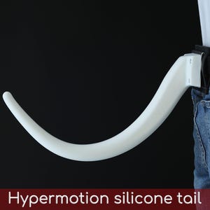 May include: A white, curved silicone tail attached to a person's belt loop, with the text "Hypermotion silicone tail" at the bottom. The tail is against a black background, with a portion of the person's blue jeans visible.