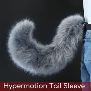 May include: A long, fluffy, grey faux fur tail accessory attached to a black belt worn over blue denim jeans. The tail has a curved, voluminous shape with dense, soft-looking fibers. The text Hypermotion Tail Sleeve is visible at the bottom.