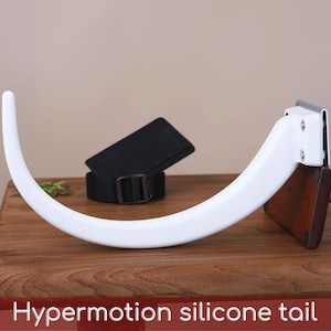 Peut inclure: Une queue en silicone blanc au design incurvé et lisse, fixée sur une base en bois foncé avec des éléments métalliques. La queue présente une forme flexible et allongée. Le texte "Hypermotion silicone tail" est inscrit en bas.
