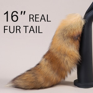 Può includere: Una coda di pelliccia vera lunga 40,6 cm. La coda è di colore marrone chiaro con una punta marrone più scura. È esposta su un supporto nero.