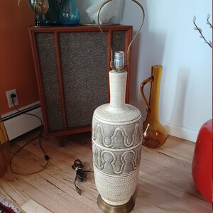May include: A white ceramic table lamp with a brown geometric pattern. The lamp has a brass base and a brass harp. The lamp is on a wooden floor.