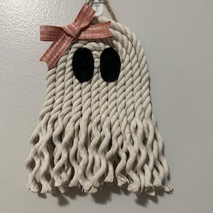 May include: A handmade Halloween ghost decoration made of off-white rope. The ghost has black oval eyes and a rust-colored bow. The ghost is hanging on a wall.