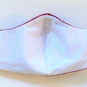 K&ouml;nnte beinhalten: Eine wei&szlig;e Baumwoll-Gesichtsmaske mit einer roten und wei&szlig;en gemusterten Au&szlig;enschicht. Die Maske hat zwei elastische Ohrschlaufen.