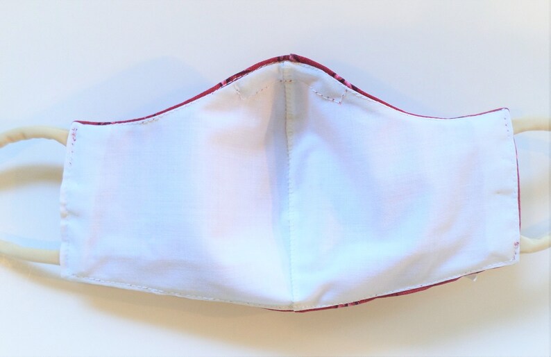 K&ouml;nnte beinhalten: Eine wei&szlig;e Baumwoll-Gesichtsmaske mit einer roten und wei&szlig;en gemusterten Au&szlig;enschicht. Die Maske hat zwei elastische Ohrschlaufen.