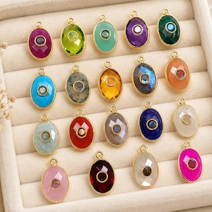 May include: A collection of twenty oval-shaped jewellery charms arranged in a grid. Each charm features a faceted gemstone set in a gold-toned metal frame with a small circular stone in the centre. The charms display a variety of colours including pink, green, blue, purple, orange, red, and white.