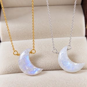 May include: Two necklaces featuring crescent moon pendants made of iridescent white stone. One necklace has a gold-toned chain, and the other has a silver-toned chain. Each pendant is faceted and displays a subtle blue sheen on the surface.