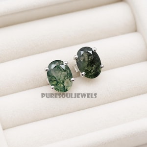 May include: A pair of oval-shaped moss agate stud earrings set in silver prongs. Each gemstone features translucent green hues with dark, branch-like inclusions throughout. The text PURESOULJEWELS is printed in dark grey below the earrings.
