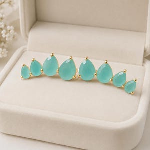 May include: An ear climber earring featuring eight faceted, teardrop-shaped light aqua stones set in gold-toned metal. The stones are arranged in a graduated size pattern, curving upward to follow the natural shape of an earlobe.
