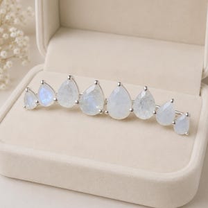 May include: A silver ear climber earring featuring eight pear-shaped moonstones arranged in a graduated arc. The stones display a translucent, milky white appearance with subtle blue iridescence, each securely held in a polished silver prong setting.