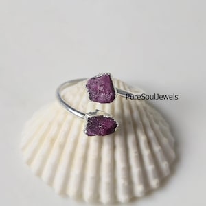 May include: A silver-toned ring with two rough-cut, deep pink gemstones. The ring is set on a white seashell, with a blurred blue object in the background. The text "PureSoulJewels" is visible on the right side of the ring.