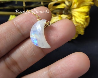 May include: A crescent moon-shaped pendant made of iridescent white moonstone with a faceted surface, suspended from a thin gold-colored metal chain. The pendant displays blue light reflections and is marked with the text PureSoulJewels.