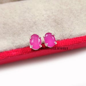 Op de afbeelding: Een paar ovale, felroze edelsteen oorknopjes met zilveren zettingen. De oorbellen worden gepresenteerd op een rode en grijze ondergrond. De tekst "PURESOULJEWELS" is zichtbaar.