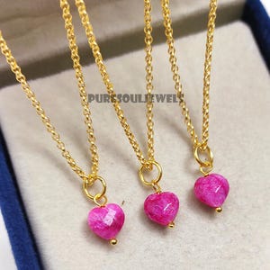 Può includere: Tre collane dorate, ciascuna con un piccolo ciondolo rosa a forma di cuore. I ciondoli hanno una texture sfaccettata e una piccola perla dorata. Le collane sono esposte su una superficie blu. Il testo "PURESOULJEWELS" è visibile.