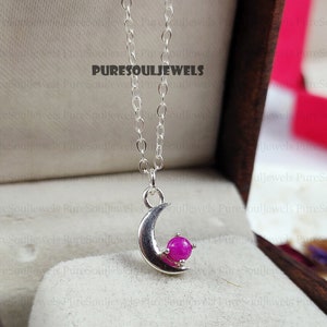 May include: A silver necklace featuring a crescent moon pendant with a magenta gemstone. The pendant hangs from a fine chain. The text "PURESOULJEWELS" is visible. The necklace is presented in a jewelry box.