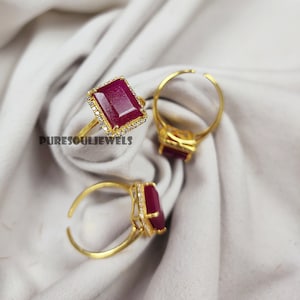 May include: Three gold rings with red gemstone settings. Each ring features a rectangular red gemstone surrounded by a halo of small clear stones. The rings are on a white fabric background.