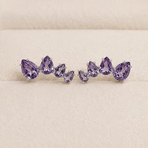 May include: A pair of silver ear climbers featuring a curved row of four pear-cut purple amethyst gemstones in each piece. The stones are arranged in graduated sizes, set in polished silver prongs, creating a fan-like shape that follows the earlobe.