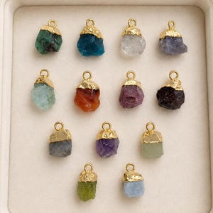 以下が含まれることがあります： 13個の原石ペンダントのコレクション。それぞれに金色の金属キャップと小さな円形のバチカンが付いています。緑、青、透明、紫、オレンジ、濃い茶色など、さまざまな色の石がトレイに並べられています。