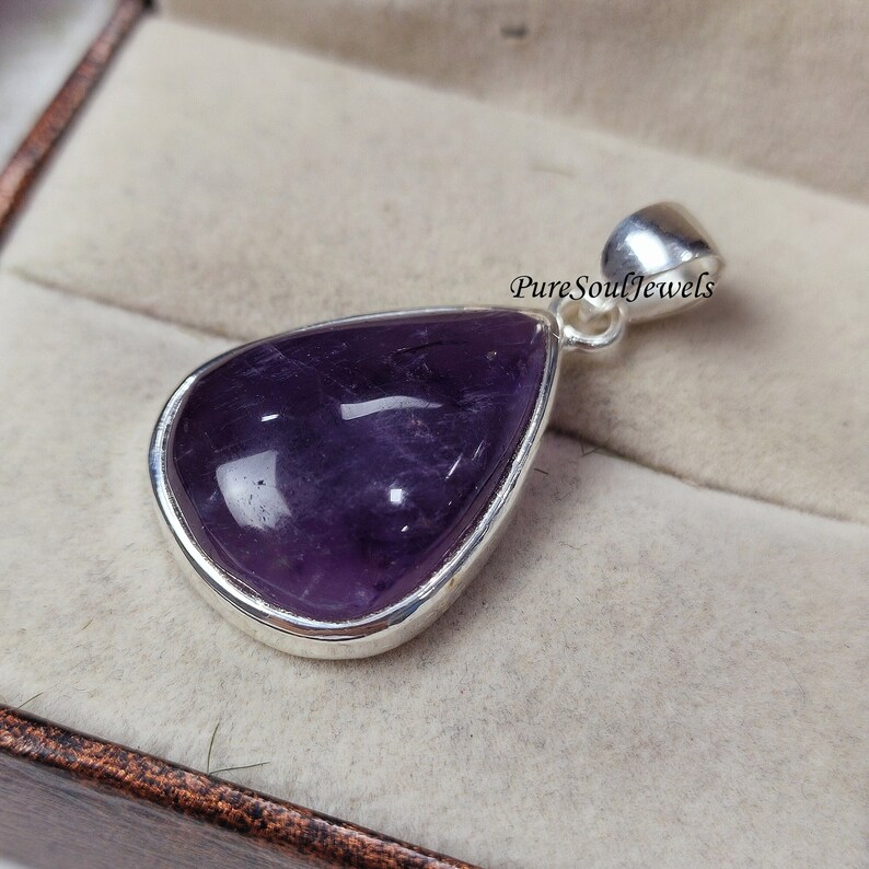 May include: A teardrop-shaped pendant with a polished, deep purple amethyst stone set in a silver bezel. The pendant has a silver bail for a chain. The text "PureSoulJewels" is visible on the right side of the pendant.