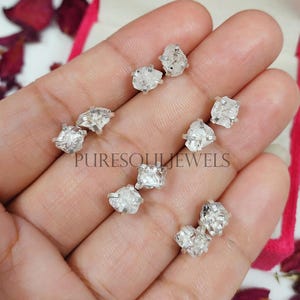 Puede incluir: Primer plano de una mano sosteniendo un par de pendientes de diamante en bruto. Los pendientes son de plata y presentan diamantes en bruto sin cortar. Los diamantes son transparentes y brillantes.