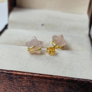 Può includere: Un paio di orecchini a forma di stella dorati con pietre di quarzo rosa. Gli orecchini sono in una piccola scatola con un esterno in pelle marrone.