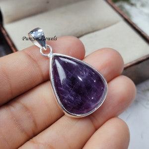 May include: A teardrop-shaped amethyst pendant with a silver-colored bezel. The polished purple gemstone has white striations and is suspended from a silver loop. The text "PureSoulJewels" is visible on the pendant's loop.