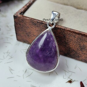 May include: A teardrop-shaped amethyst pendant with a polished, deep purple stone set in a silver-colored metal frame. The pendant has a loop at the top for a chain. The background includes a wooden box and the text "Pure Soul Jewels".