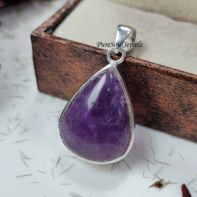 May include: A teardrop-shaped amethyst pendant with a polished, deep purple stone set in a silver-colored metal frame. The pendant has a loop at the top for a chain. The background includes a wooden box and the text "Pure Soul Jewels".