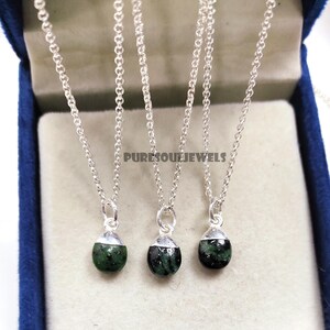 Puede incluir: Tres collares de plata con pequeños colgantes en forma de lágrima. Los colgantes son de color verde oscuro con vetas negras. Los collares se presentan en una caja de joyería azul. El texto "PURESOULJEWELS" es visible.