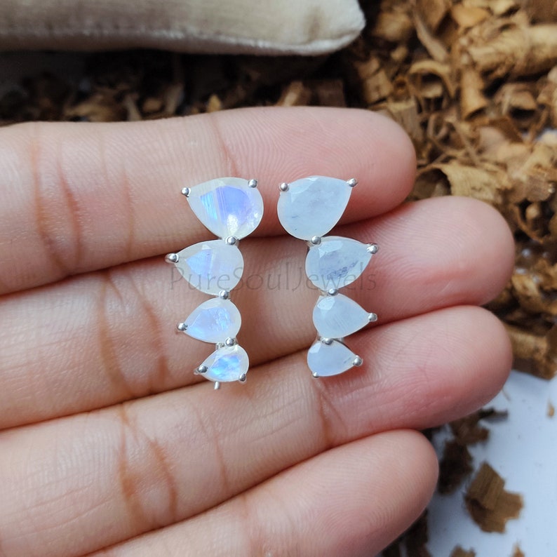 May include: A pair of silver ear climbers featuring four pear-shaped moonstones each, arranged in a cascading vertical line. The stones exhibit a translucent white appearance with subtle blue iridescence. The text PureSoulJewels is visible.