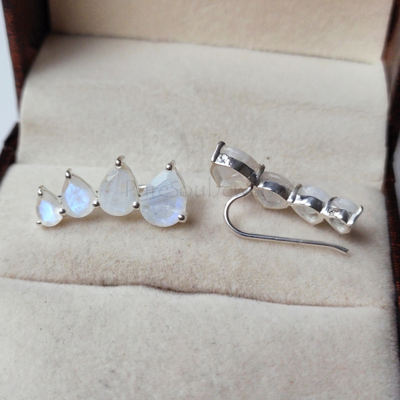 May include: A pair of silver ear climbers featuring four pear-shaped moonstones in each piece. The stones are arranged in a graduated size pattern, showing iridescent blue flashes. The metal settings are polished silver with a curved wire backing.
