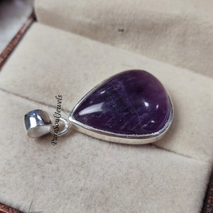 May include: A teardrop-shaped pendant with a polished, deep purple gemstone set in a silver bezel. The pendant has a silver bail for a chain. The text "Pure Soul Jewels" is printed on the side. Jewelry.