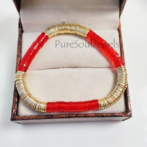 May include: A red and gold bracelet featuring a stack of gold rings and red beads. The bracelet is displayed in a small box with a white interior and a brown exterior. The text "PureSoulJewels" is visible on the box.
