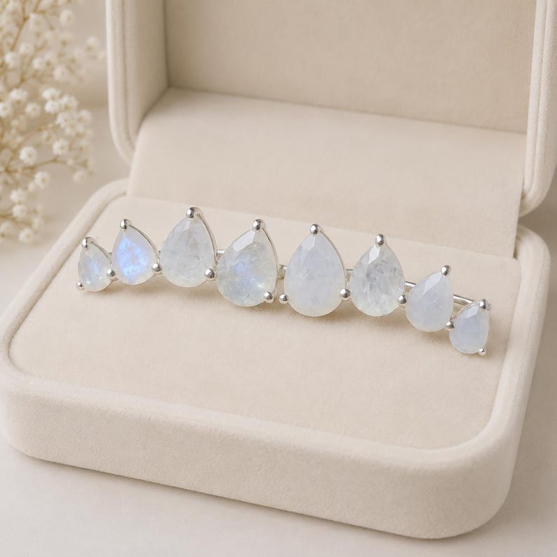 May include: A silver ear climber earring featuring eight pear-shaped moonstones arranged in a graduated arc. The stones display a translucent, milky white appearance with subtle blue iridescence, each securely held in a polished silver prong setting.