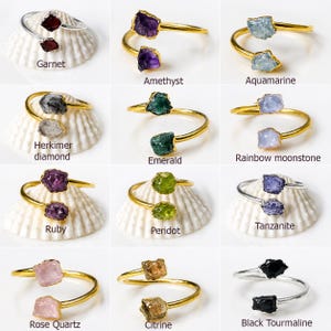 Op de afbeelding: Een verzameling edelsteenringen, elk met een unieke ruwe steen. De ringen hebben een gouden of zilveren band, met stenen zoals granaat, amethist, aquamarijn, Herkimer diamant, smaragd, regenboog maansteen, robijn, peridot, rozenkwarts, citrien en zwarte toermalijn.