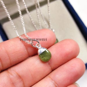 以下が含まれることがあります： 小さなオリーブグリーンの宝石ペンダントが付いたシルバーネックレス。ペンダントは涙の形をしており、シルバーのキャップが付いています。チェーンは繊細で、ペンダントは手で持たれています。「Puresouljewels」というテキストが表示されています。