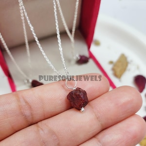 May include: A silver chain necklace with a small, rough, red gemstone pendant. The pendant is attached to the chain with a silver loop and a silver bead.