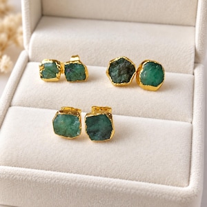 May include: Three pairs of stud earrings featuring raw, uncut green emerald stones with irregular shapes. Each stone is encased in a bright gold-toned metal setting with a textured, organic edge, showcasing the natural variations of the gems.