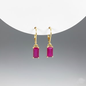 May include: A pair of gold-colored leverback earrings, each featuring a rectangular, vibrant magenta-colored gemstone. The earrings are suspended from a curved white surface against a gradient gray background. The gemstones are approximately 0.5 inches in length.