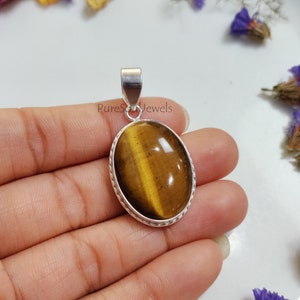 Op de afbeelding: Een zilveren hanger met een bruine en gouden tijgeroog edelsteen. De hanger heeft een gevlochten zilveren rand en is ovaalvormig. De tekst "PureSoul Jewels" is zichtbaar in de afbeelding.