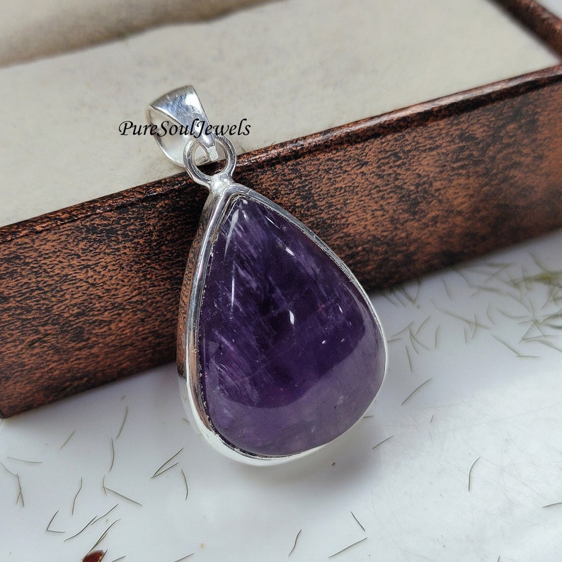 May include: A teardrop-shaped pendant with a polished, deep purple gemstone set in a silver-colored metal frame. The pendant has a bail for a chain and is displayed on a white surface. The text "Pure Soul Jewels" is visible.