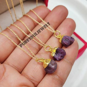 Peut inclure: Colliers dorés avec des pendentifs en pierres précieuses violettes brutes et non taillées. Les pendentifs sont sertis dans du métal doré. Les colliers sont présentés sur une main. Le texte "PURESOULJEWELS" est visible.