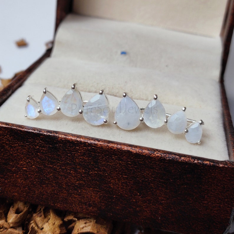 May include: A pair of silver ear climbers featuring a row of seven pear-shaped moonstones in varying sizes. Each gemstone is set in a prong setting, displaying a translucent white appearance with subtle blue iridescence. The text PureSoulJewels is visible.
