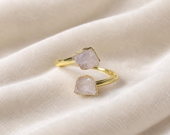 May include: A gold-toned open ring featuring two raw, pale pink crystal stones at each end. The band is smooth and polished, while the stones have a natural, irregular, and textured appearance. The design is adjustable with a gap between the stones.
