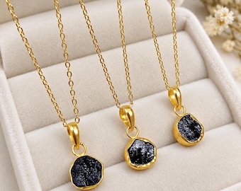 Peut inclure: Trois colliers dorés, chacun doté d'un petit pendentif en pierre noire brute et irrégulière, serti dans une monture dorée polie. Les pendentifs sont suspendus à de fines chaînes à maillons dorés, créant un contraste entre la texture sombre des pierres et la finition métallique brillante et lisse des montures et des chaînes.