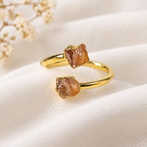 May include: A gold-toned open ring featuring two raw, amber-colored crystals at each end. The band is smooth and polished, contrasting with the textured, irregular surfaces of the two stones. The design is adjustable, with the ends not meeting.